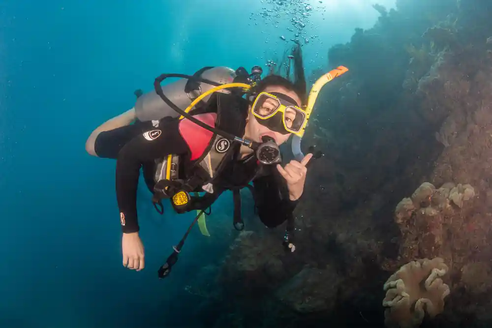A diver in a hood giving a shaka sign over the reef
