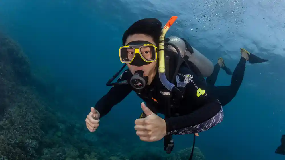 A diver in a hood giving a thumbs up beside the coral