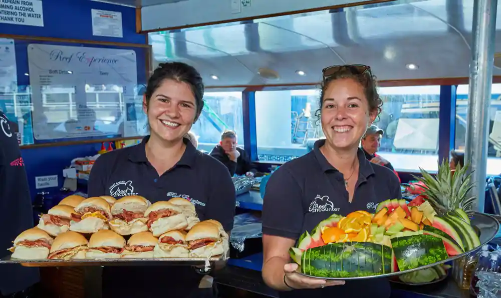 Two crew holding out trays of filled rolls and a cut fruit platter
